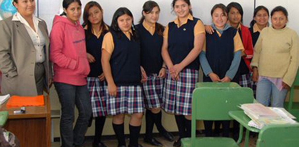 Eine Mädchengruppe in Schuluniform steht in einer Klasse neben ihrer Lehrerin. Rechts sind zwei grüne Stühle mit Schulsachen.