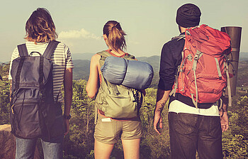 Drei junge Menschen stehen mit Wandergepäck auf einer Anhöhe und sehen in die Ferne. 