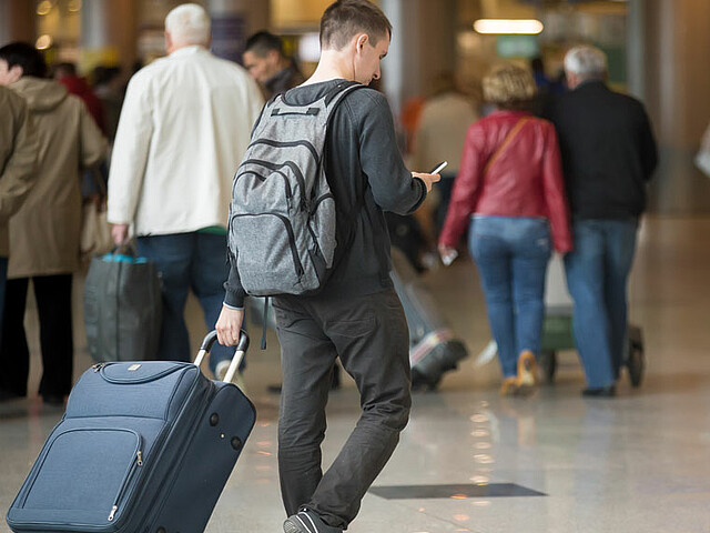 Ein junger Mann ist mit Rucksack und Koffer bepackt und schaut auf sein Handy, das er in der rechten Hand hält, während er in einem Flughafengebäude nach vorne läuft.